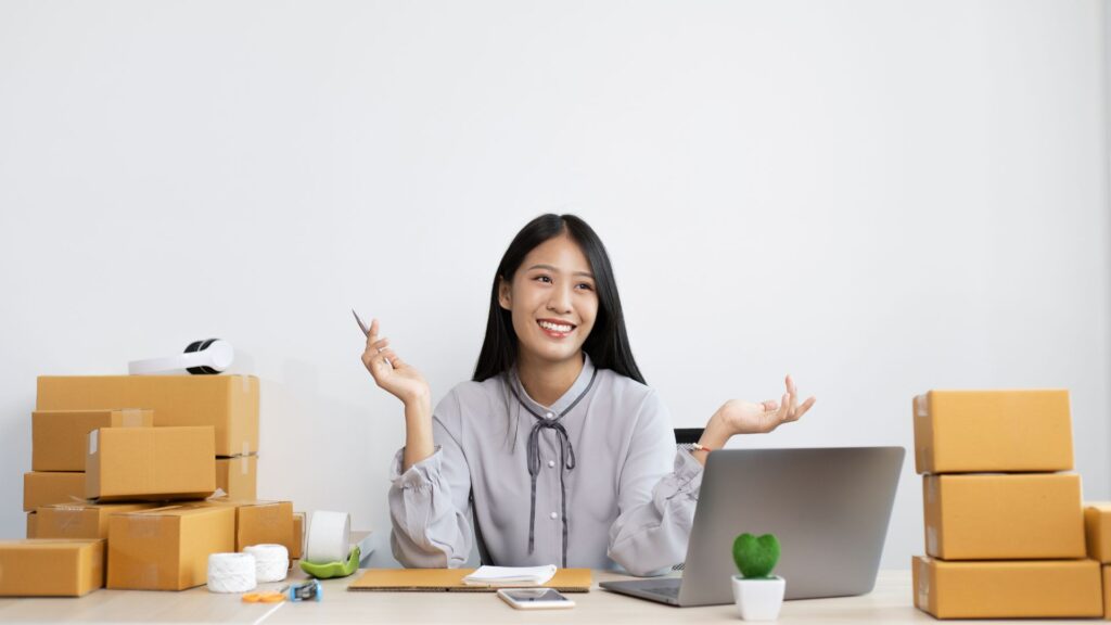 Mujer sonriente con laptop y cajas de envío representando habilidades digitales para emprender online.